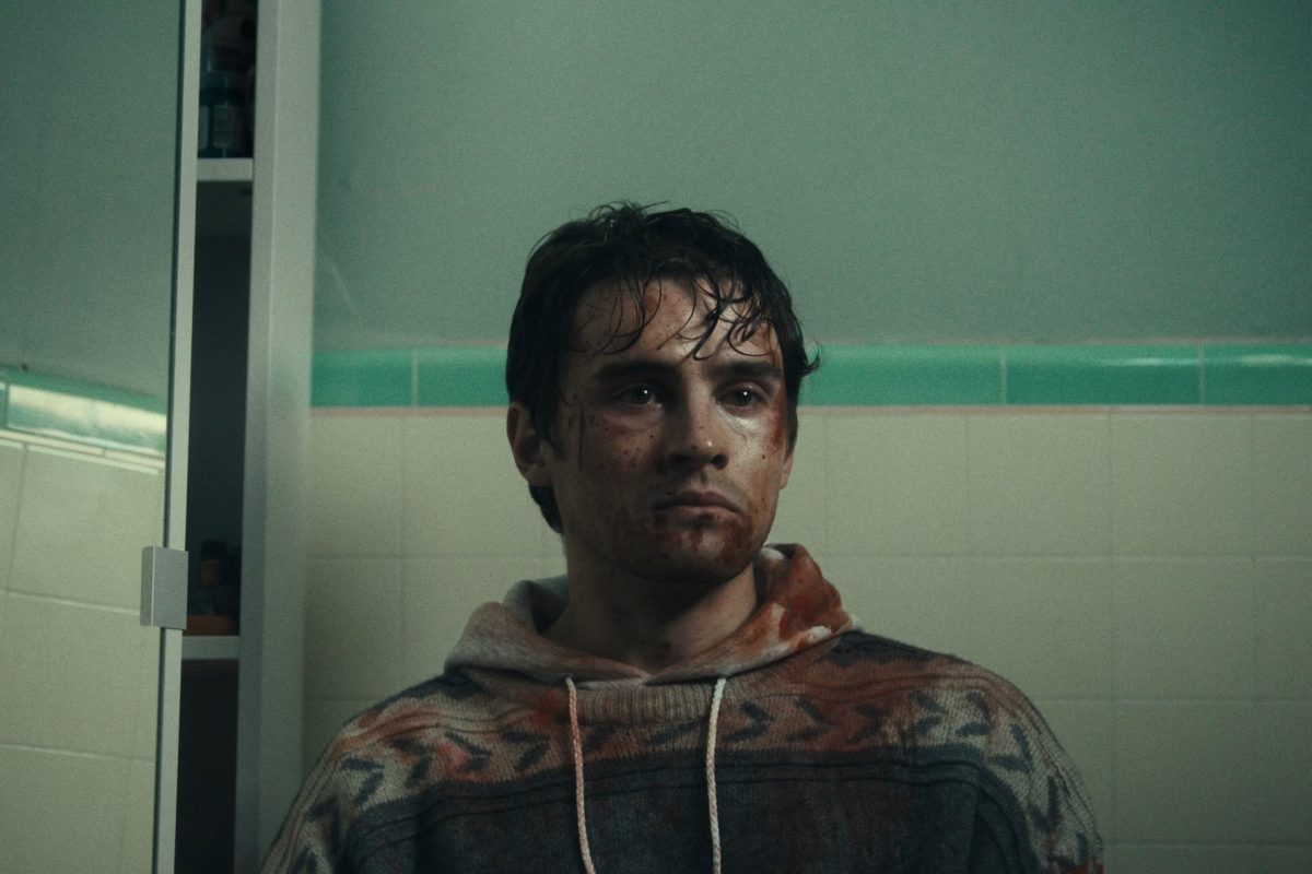 A man stands in a bathroom. He has a blank expression, and his clothes and face are covered in blood.